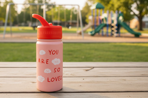 Load image 1 in gallery view. Pink water bottle with red lid and 'You are so loved' text on picnic table