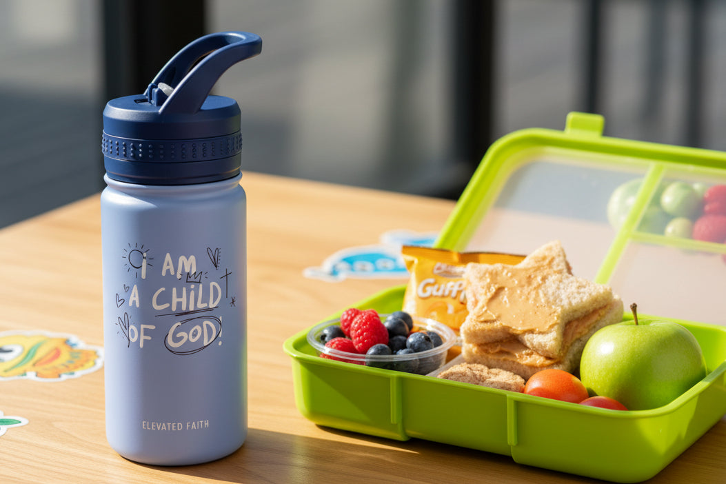 Blue water bottle with 'I am a child of God' text next to a green lunchbox with snacks on a wooden table